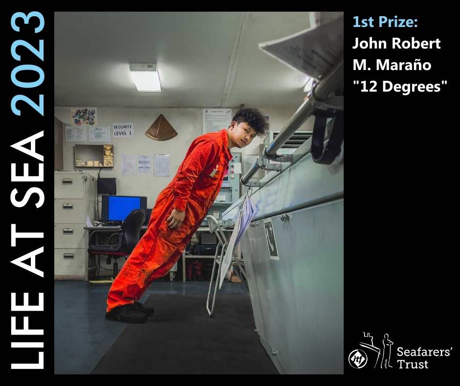 A graphic showing the Ist Prize photo on a black background with text reading Life At Sea 2023 1st Prize Winner John Robert M. Marano, "12 Degrees", the photo shows a seafarer standing on the ships' bridge apparently at an impossible angle in relation to the floor, however the clinometer on the wall behind shows it is actually the ship that is at a steep angle.