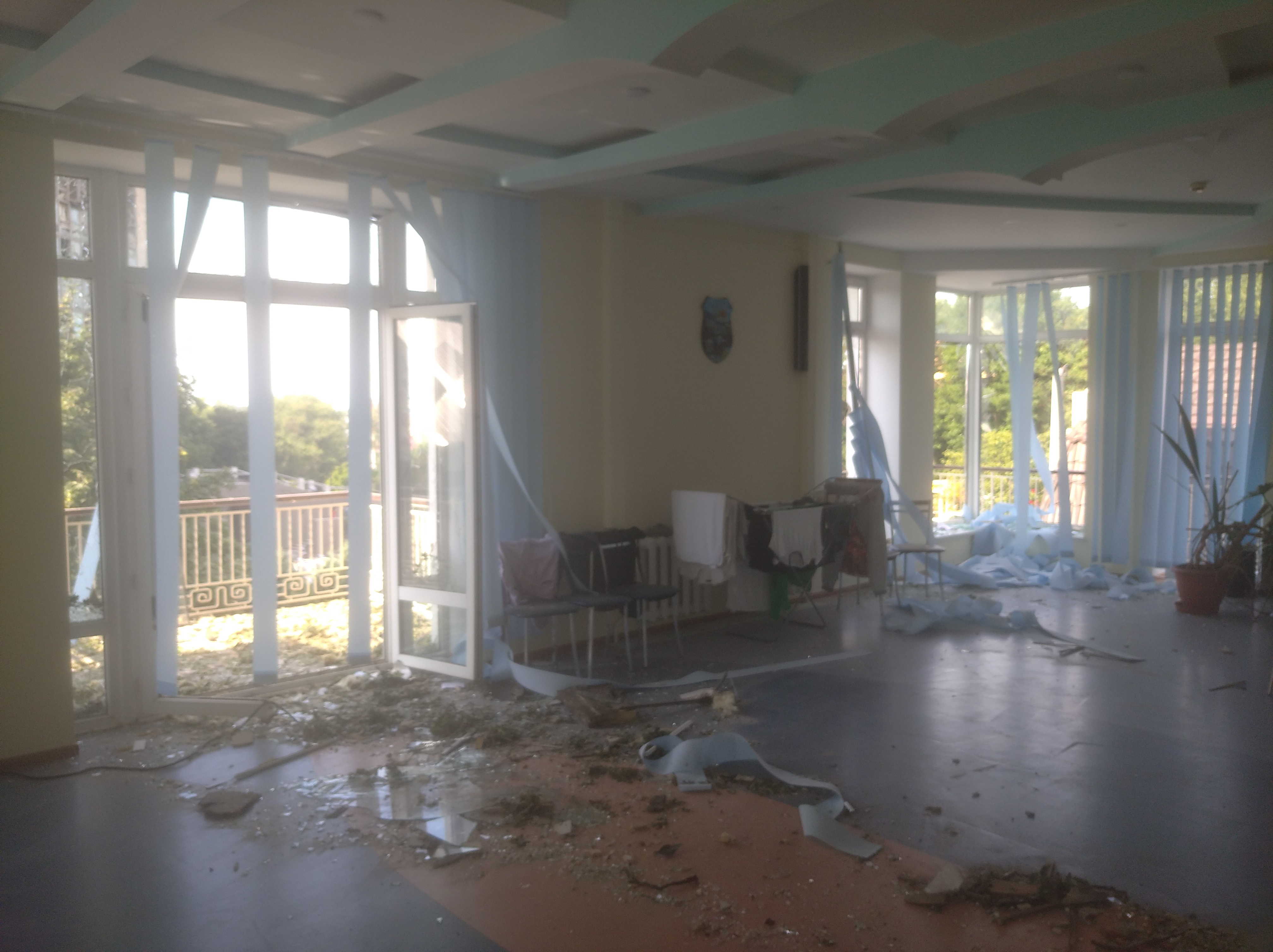 A picture of a large open room with the windows broken by gunfire and smashed glass and debris on the hardwood flooring