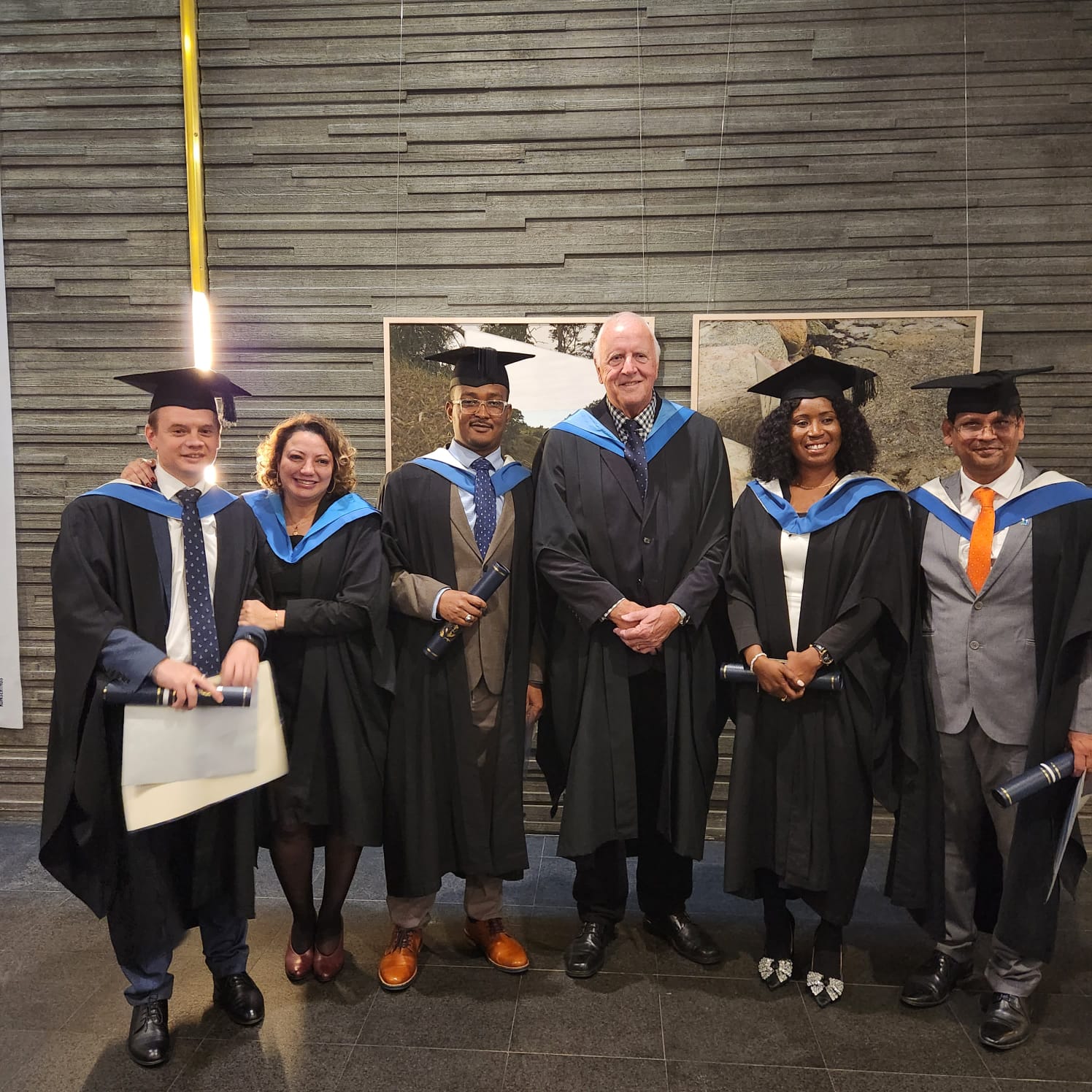 Honorary Fellow, Brian Orrell and Graduating students Mr Lamir Ado Mohammed, Ms. Regina Chokolay Conteh-Khali, Mr Pankaj Kumar, Ms. Mariami Kakabadze and Mr Sergii Kazantsev, wearing official graduating robes, standing in front of a WMU banner 